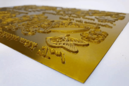 A close-up view of a yellow etched photopolymer plate featuring raised designs of butterflies, text, and decorative elements on its surface.